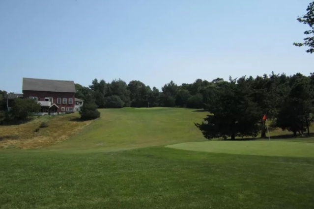 Cotuit High Ground Golf Club Bog Course green with red flag, sloping fairway, trees, and house under clear sky