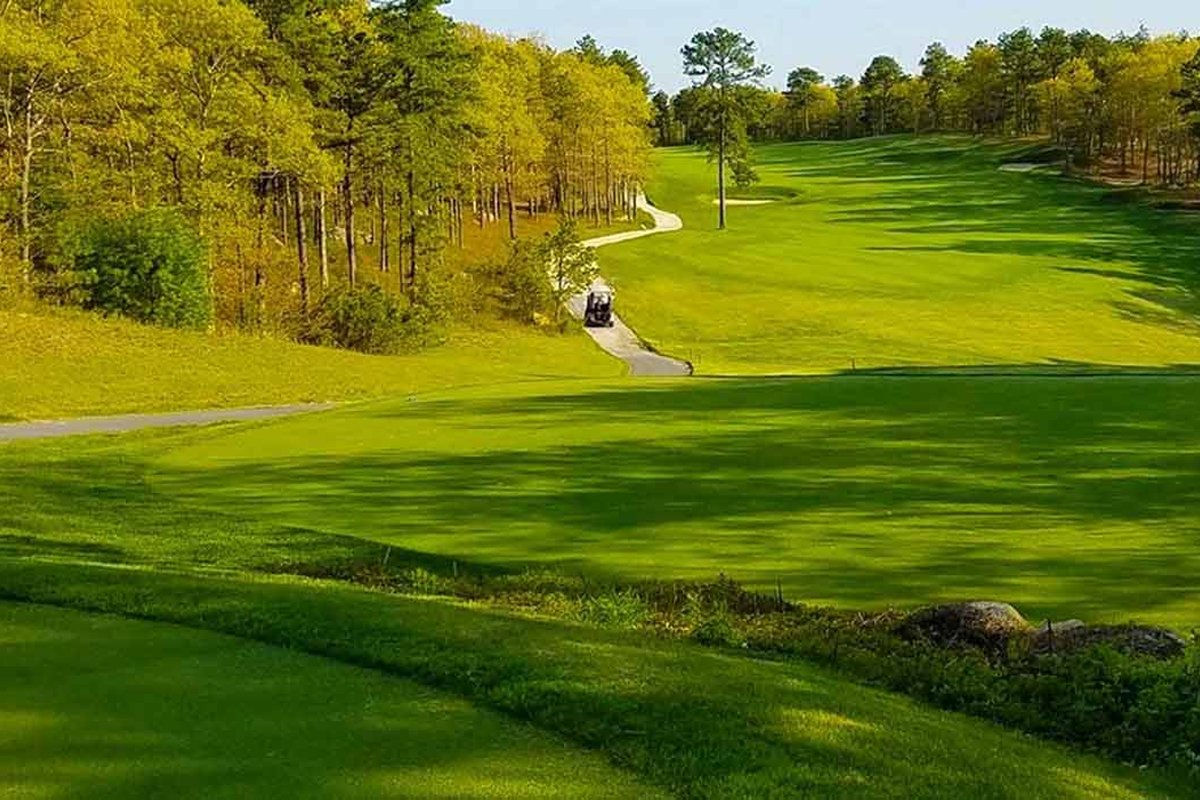 Rolling fairway and putting green at Crosswinds Golf Club Ouimet course in Plymouth, MA, bordered by tall trees under clear sky