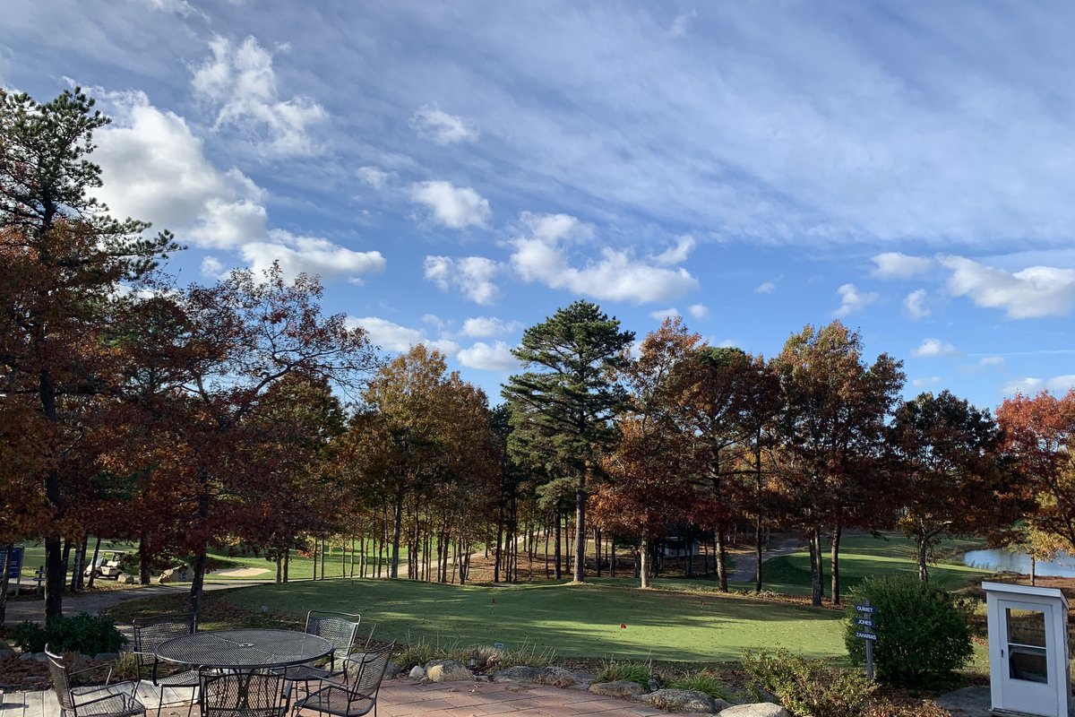 Brick patio with empty metal chairs overlooks the Zaharias course at Crosswinds Golf Club, featuring trees, green fairway, and blue sky with clouds