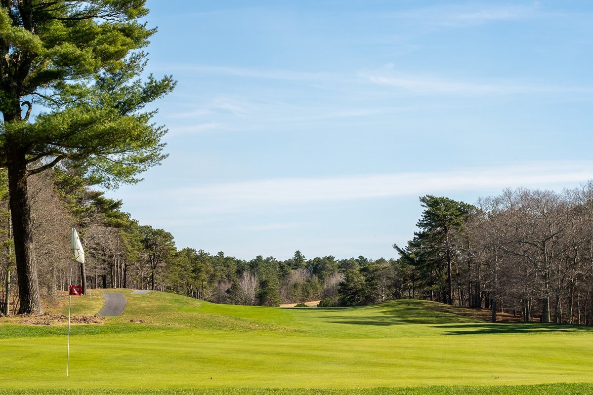 Flag on manicured green at Atlantic Country Club, Plymouth, MA, fairway lined with pine and bare trees under blue sky