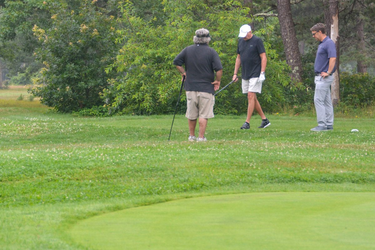 Three golfers stand beside the green at Falcon Golf Course in Sandwich, MA, surrounded by trees and lush rough grass.