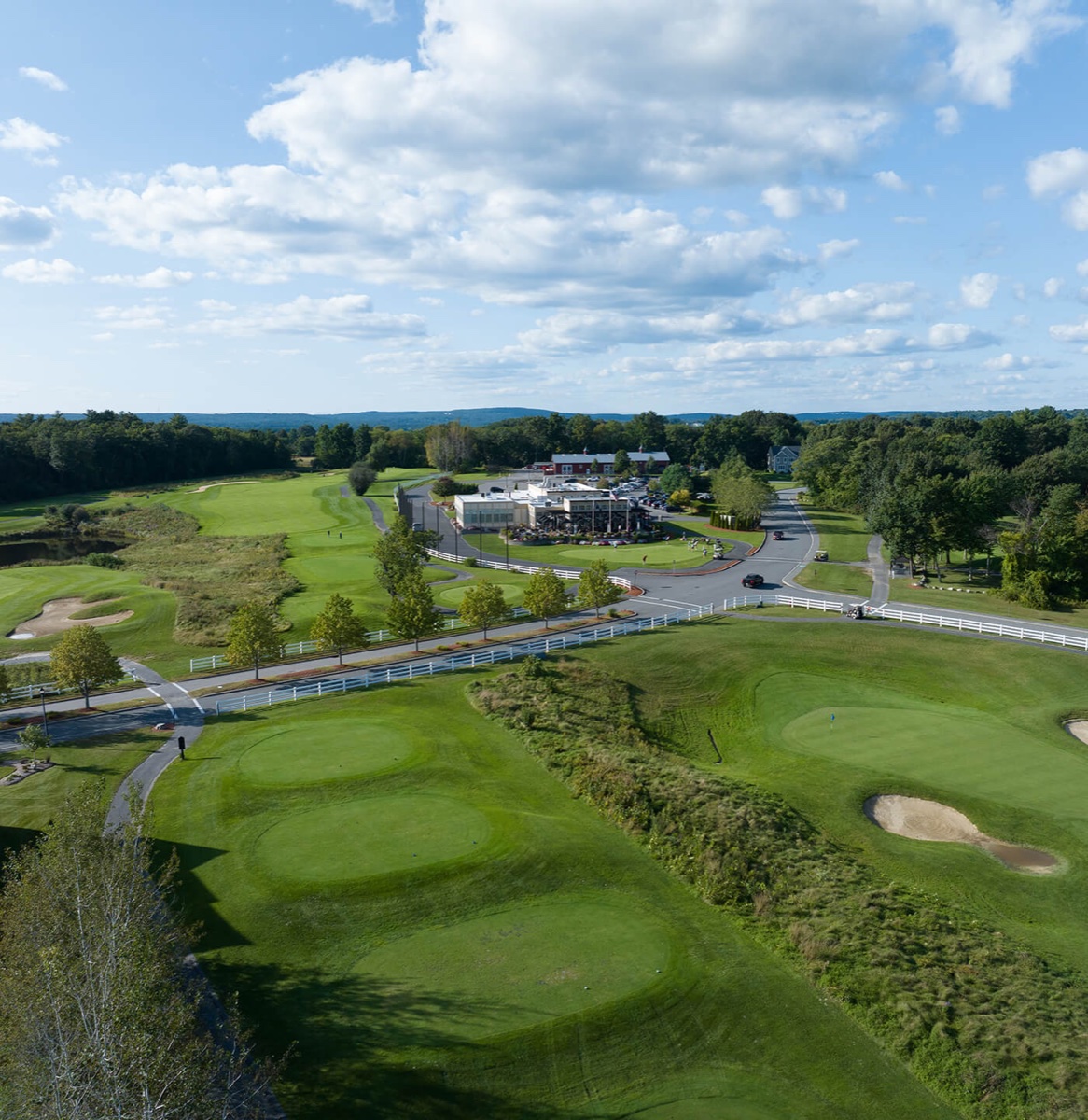 Aerial view of Four Oaks Golf Course in Dracut, MA featuring lush fairways, greens, bunkers, clubhouse, trees, and partly cloudy sky