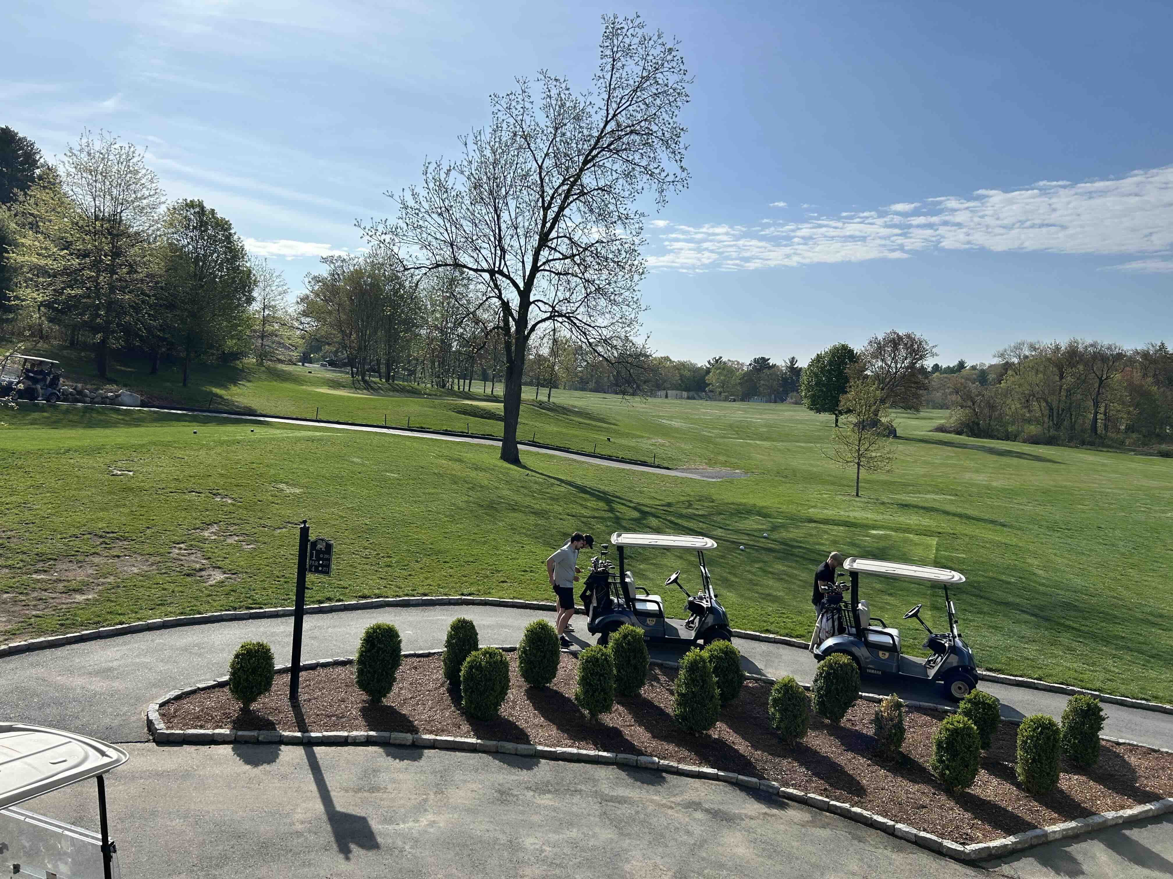 Golfers prepare near golf carts on a paved path with lush fairways, scattered trees, and blue sky at Foxborough Country Club.