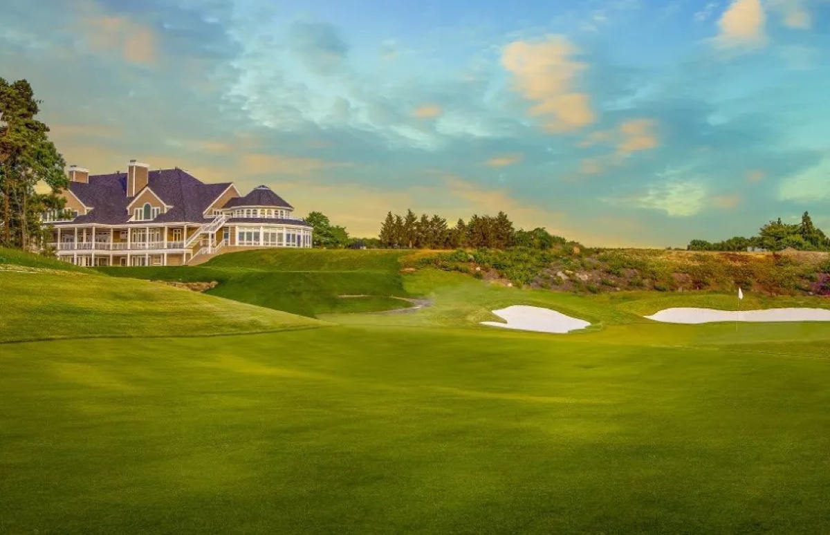 Expansive green fairway at The Cape Club in Falmouth, MA with sand bunkers, clubhouse, trees, and a colorful sunset sky