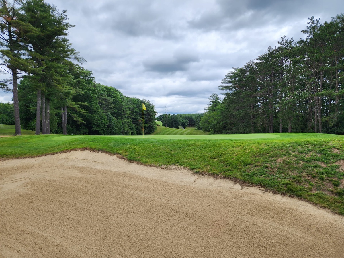 Sand bunker in the foreground with green, yellow flag, lush fairway, and dense trees at Gardner Municipal GC, cloudy sky above