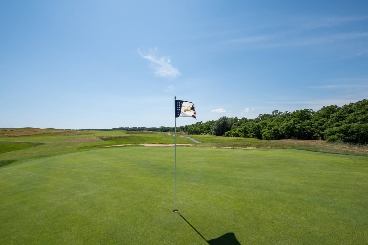 American flag pin stands on a well-manicured green at Bass River Golf Course, with fairways and trees under clear blue sky