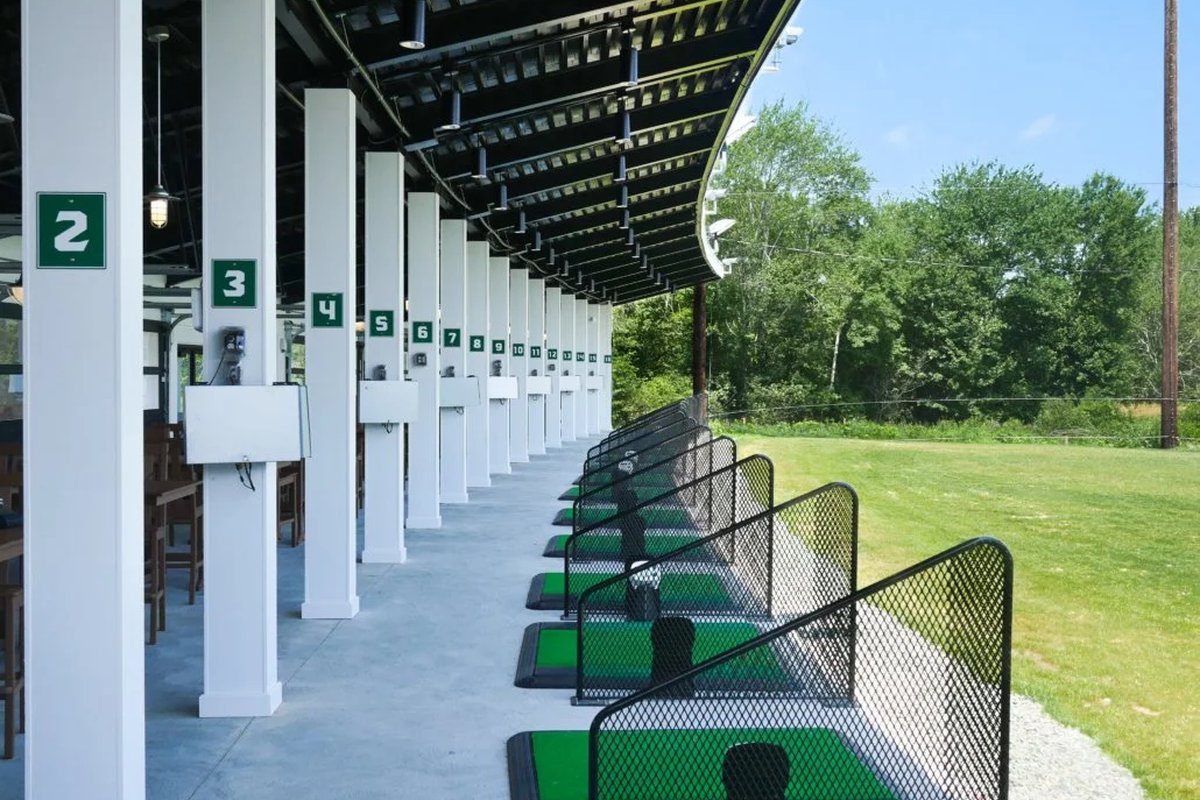 Covered driving range at Green Harbor Golf Club with numbered stalls, green mats, white pillars, and trees in the background