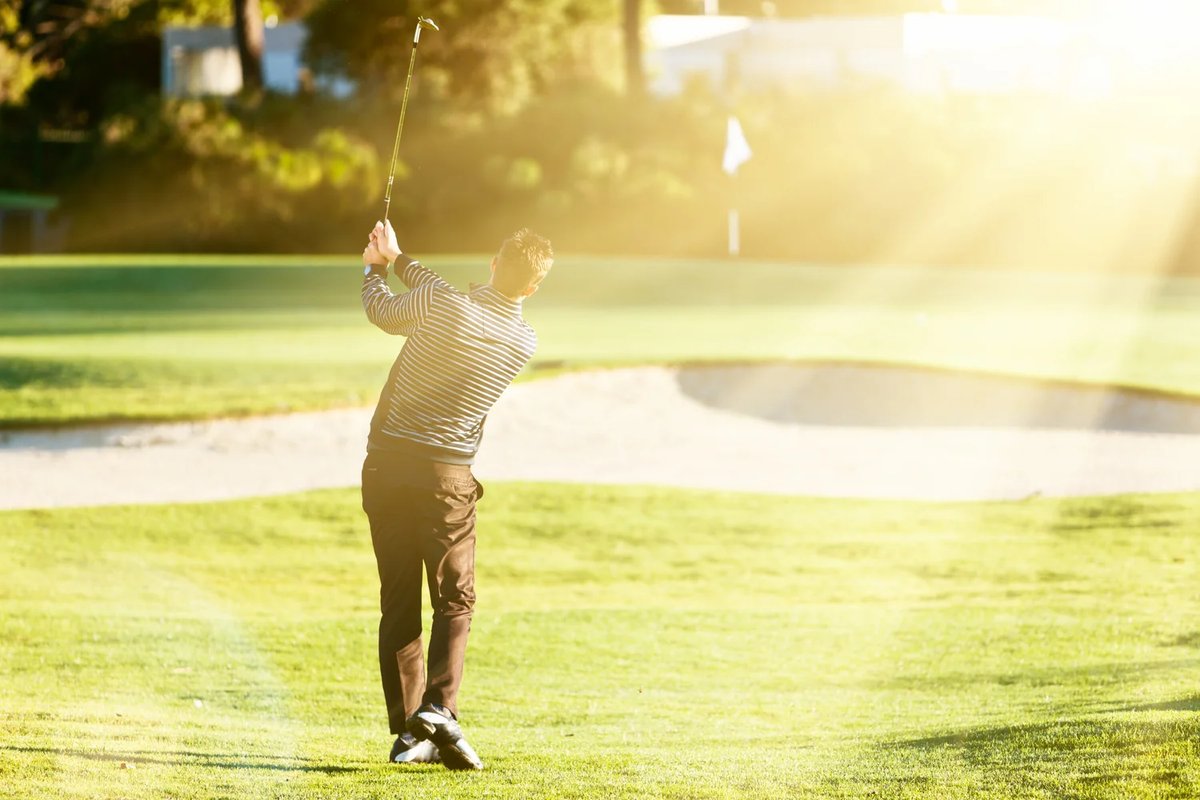 Golfer in striped shirt hitting iron shot toward sunlit green and pin at Groton Country Club, sand bunker in background