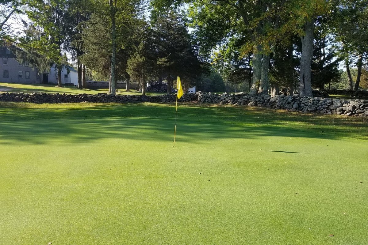 Bright green putting green with yellow flagstick at Hemlock Ridge Golf Course, bordered by stone wall and tall trees under clear sky