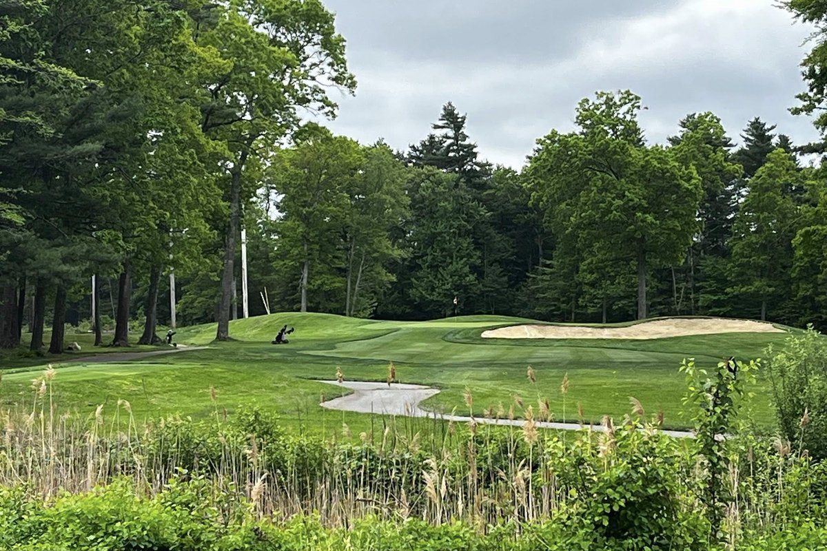 Lush green fairway and putting green at The Back Nine Club in Lakeville, MA, bordered by trees and a large sand bunker under cloudy skies