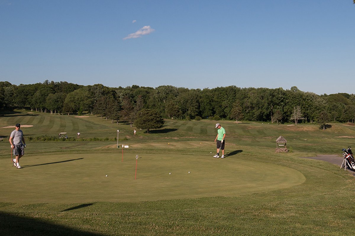 Two golfers practice putting on a green at Hillside Country Club, lush fairways and trees under a clear blue sky