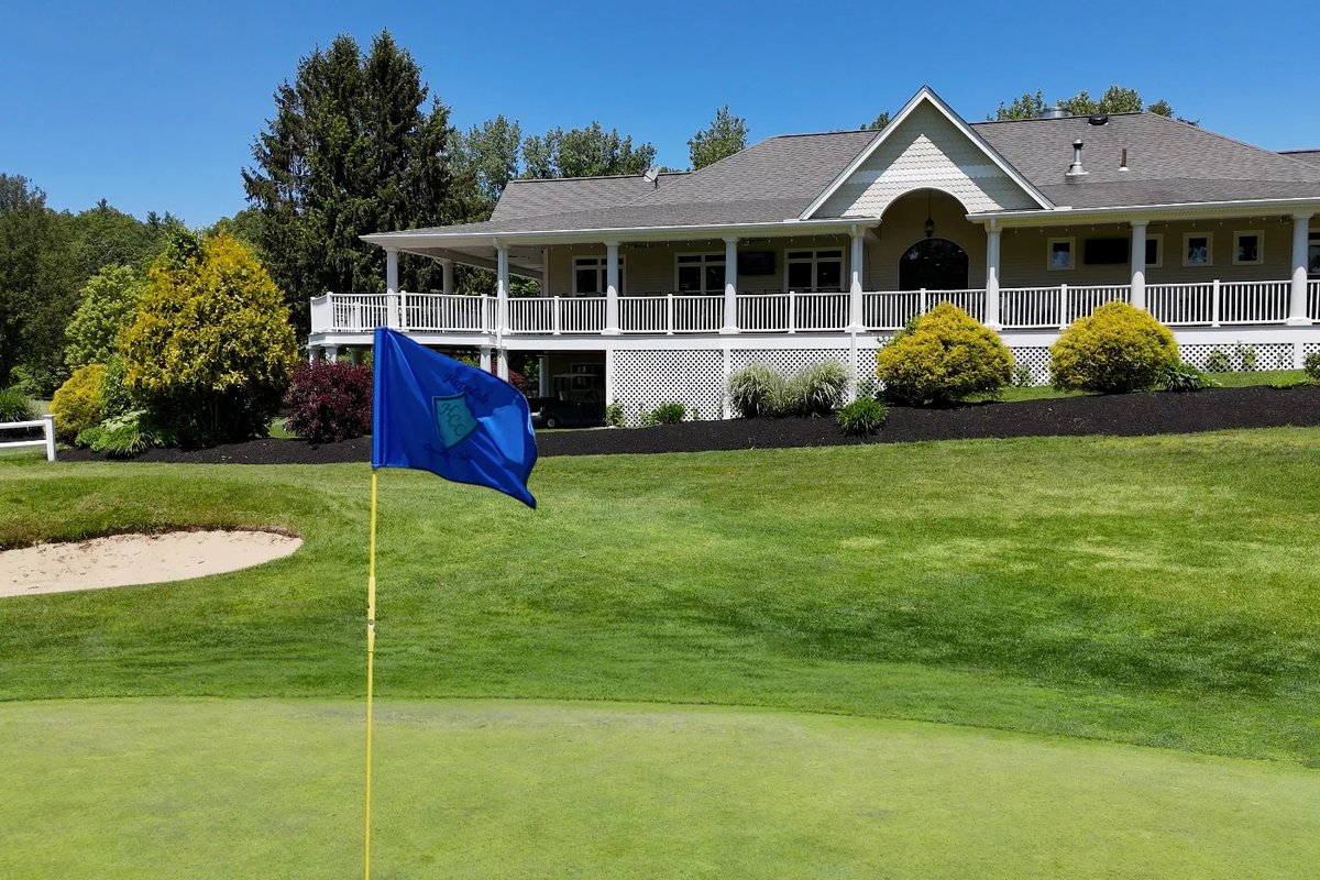 Blue pin flag on manicured green near sand bunker, with Hopedale CC clubhouse and lush landscaping in background under blue sky
