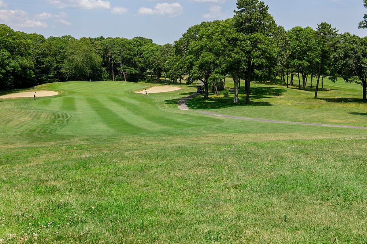 Wide fairway at Bay Pointe Club in Wareham, MA, bordered by sand bunkers, mature trees, and a blue sky with scattered clouds
