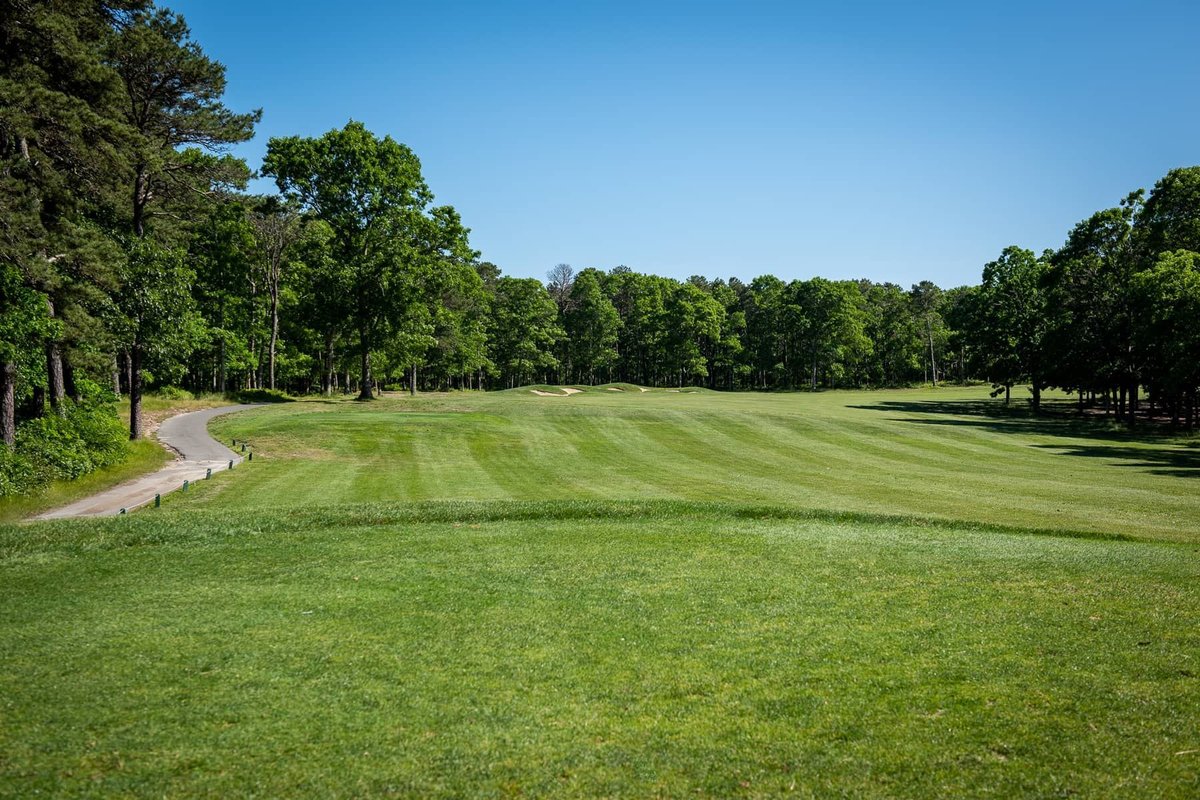 Wide fairway at Bayberry Hills Golf Club Blue/Red course with lush green grass, tree-lined edges, sand bunkers, and clear blue sky