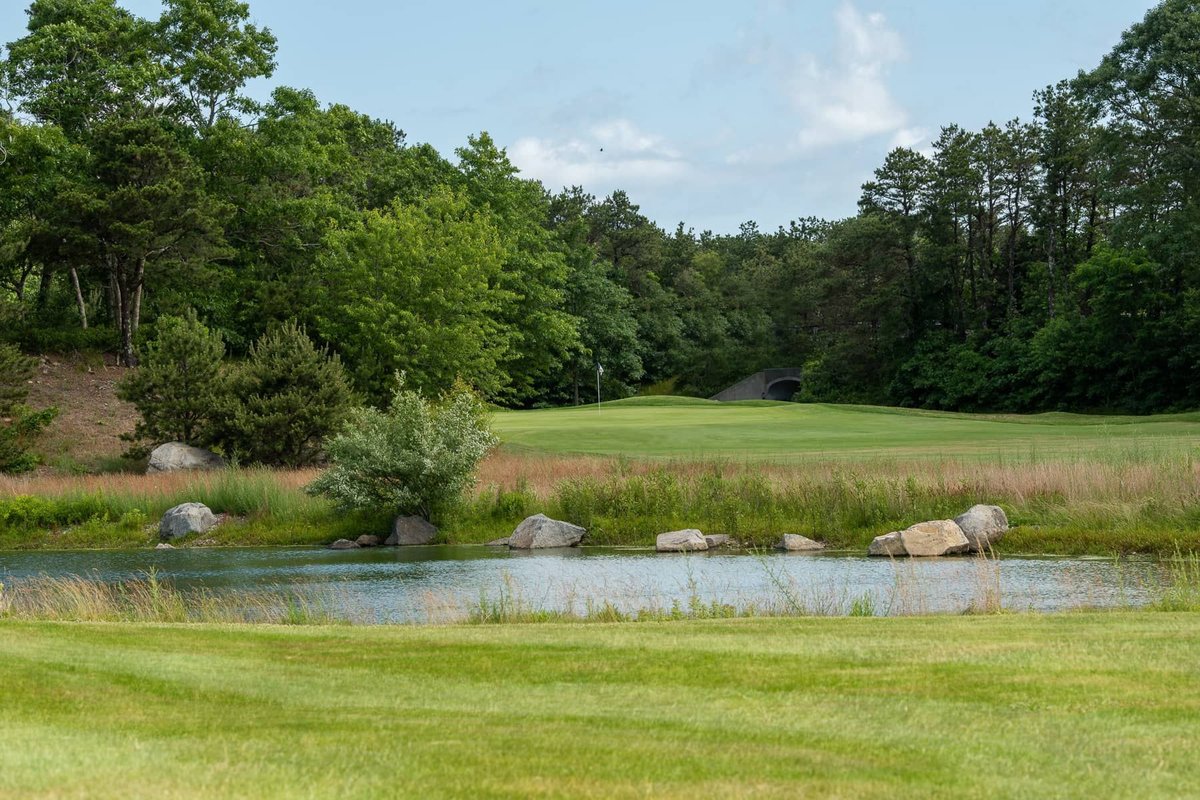 Lush fairway at Bayberry Hills Golf Club Red/White course in Yarmouth, MA with pond, rocks, and dense trees under blue sky