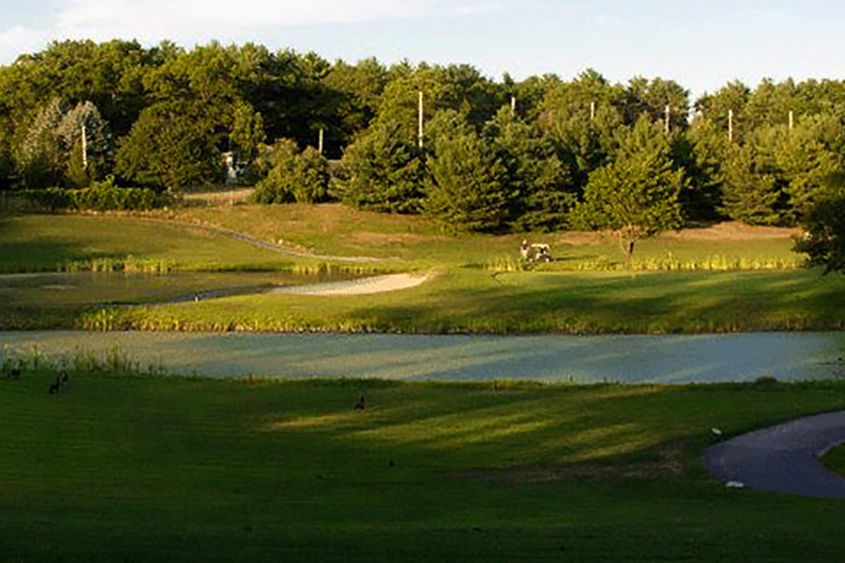 Tree-lined fairway and putting green at Lakeville Country Club, with a small pond, golf cart, sand bunker, and blue sky