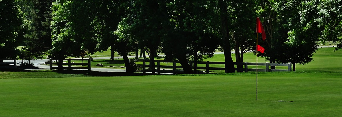 Lush green putting green with red flagstick at Little Harbor Country Club, shaded by tall trees and bordered by a white fence