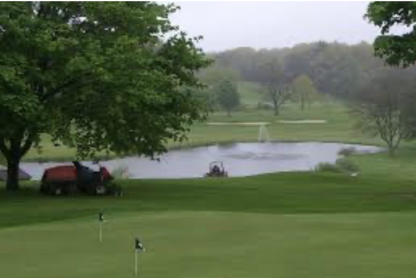 Greens with putting flags in foreground, maintenance vehicles near a tree, small pond and lush fairways at Maynard Country Club