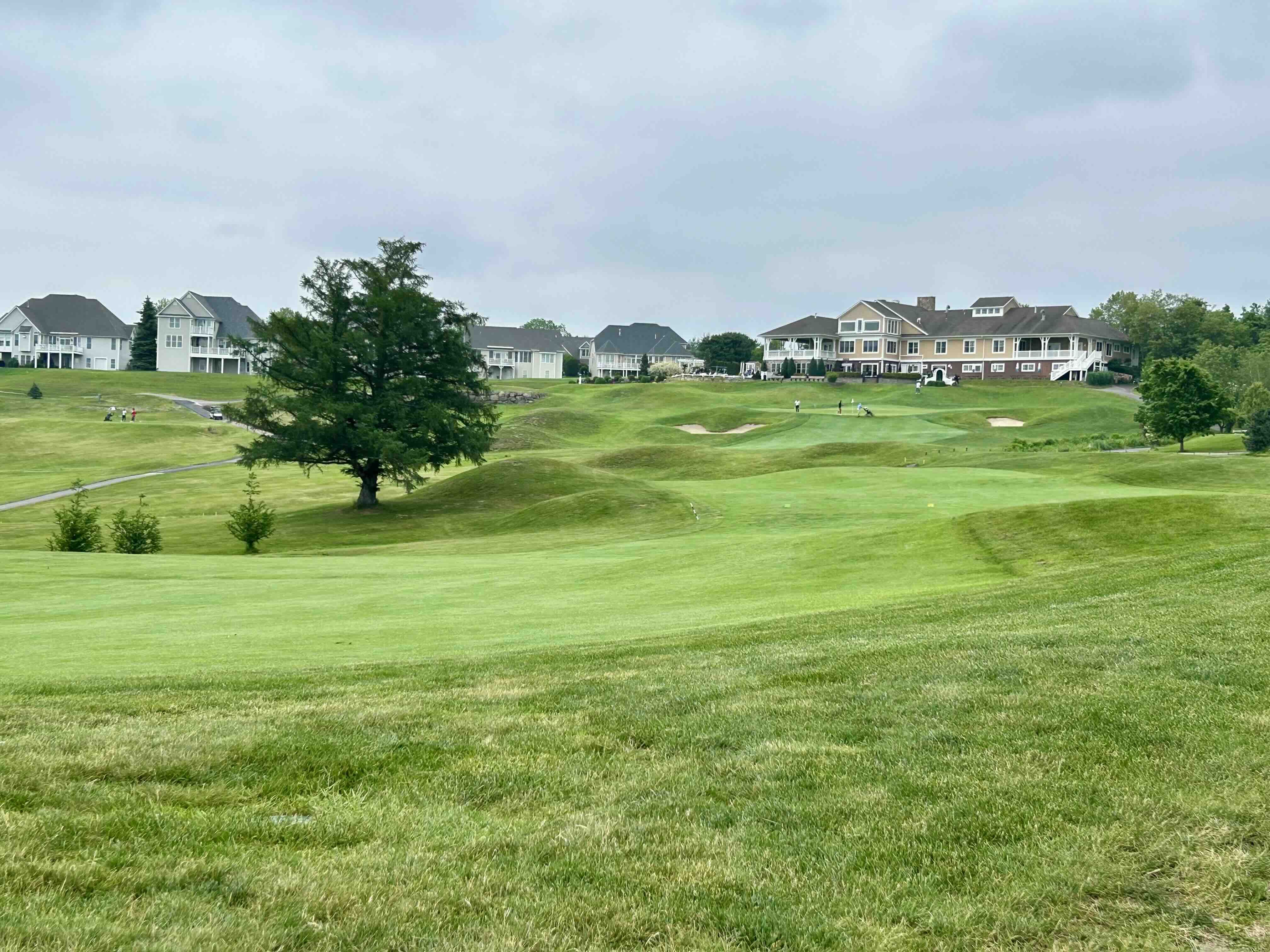 Rolling fairway at Merrimack Valley Golf Course with sand bunkers, large tree, cloudy sky, and clubhouse with homes in background