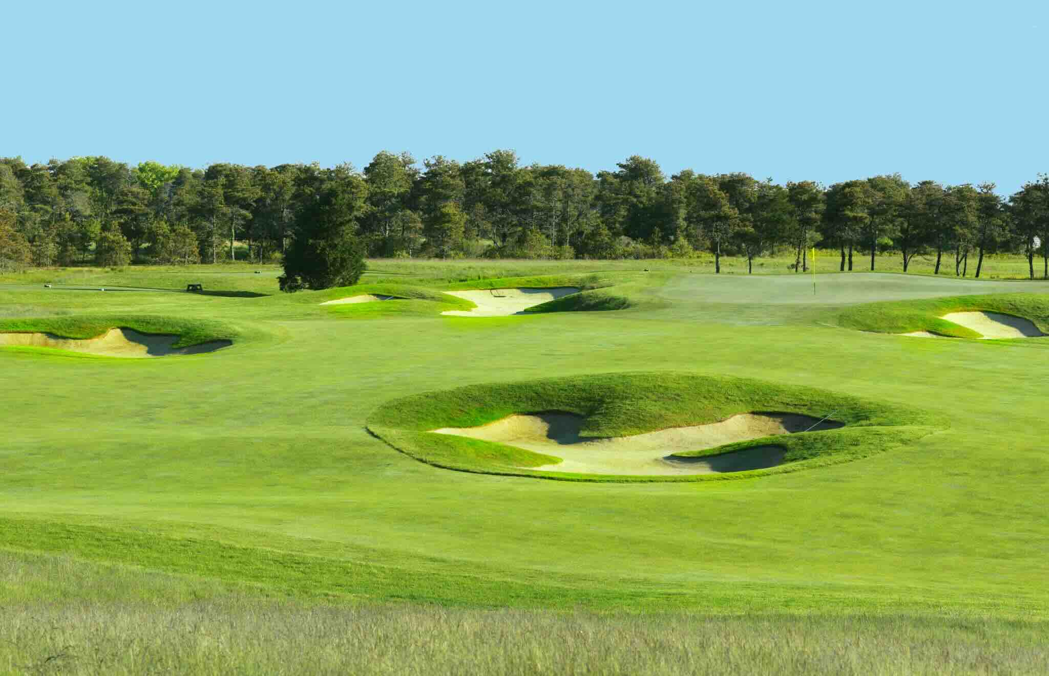 Lush fairways and multiple sand bunkers on a sunny hole at Miacomet Golf Course, with dense trees in the background and clear blue sky.