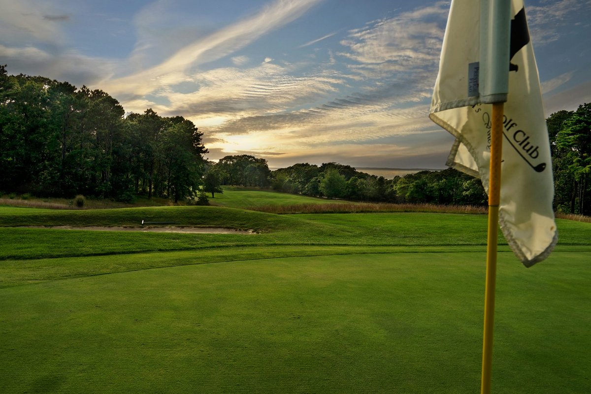 Flagstick on the green at Mink Meadows Golf Club with lush fairway, trees, and a dramatic evening sky in Tisbury, MA