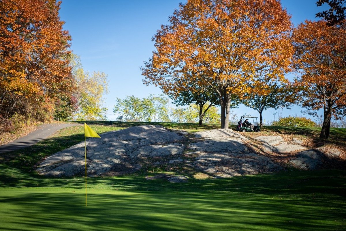Yellow flag on green at Mount Hood Golf Course in Melrose, MA, with rocky terrain, autumn trees, and blue sky in background