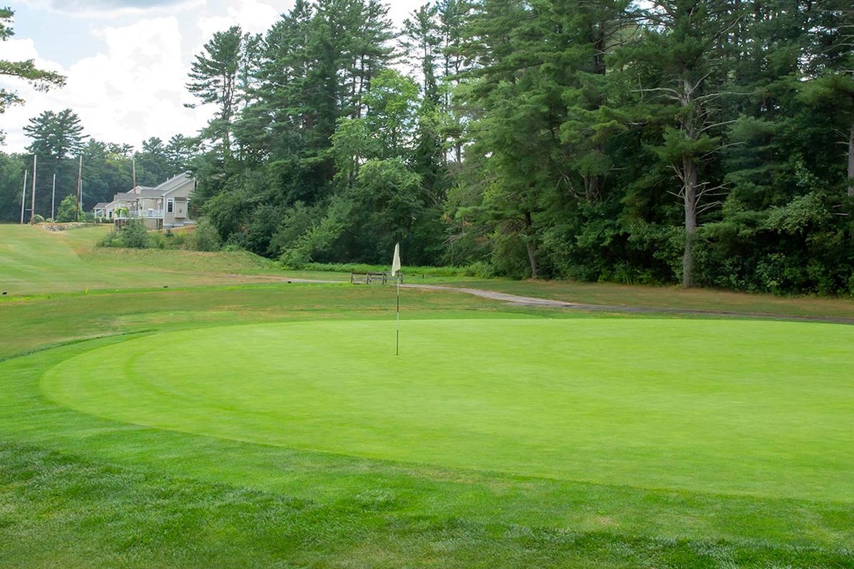 Lush green at New Meadows Golf Club in Topsfield, MA with flagstick, clubhouse, surrounding trees, and blue sky with clouds