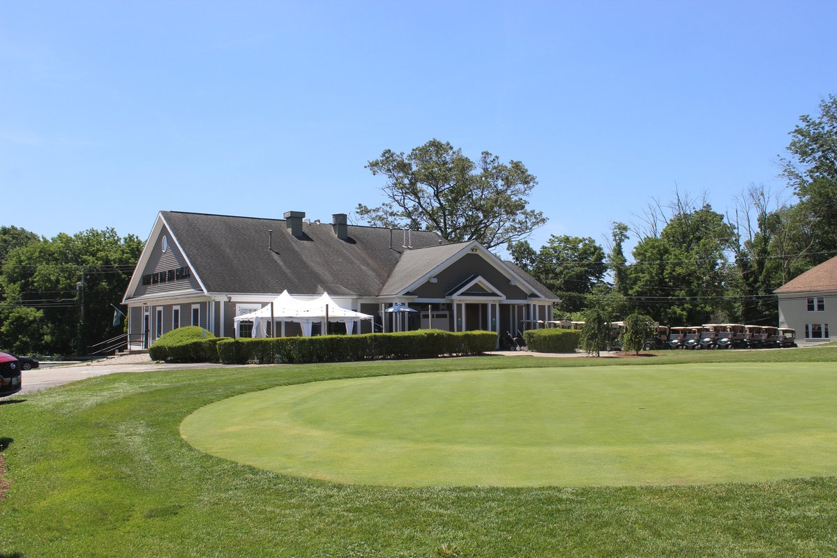 Clubhouse at Dudley Hill Golf Club with outdoor patio, golf carts lined up, lush green putting surface, and clear blue sky