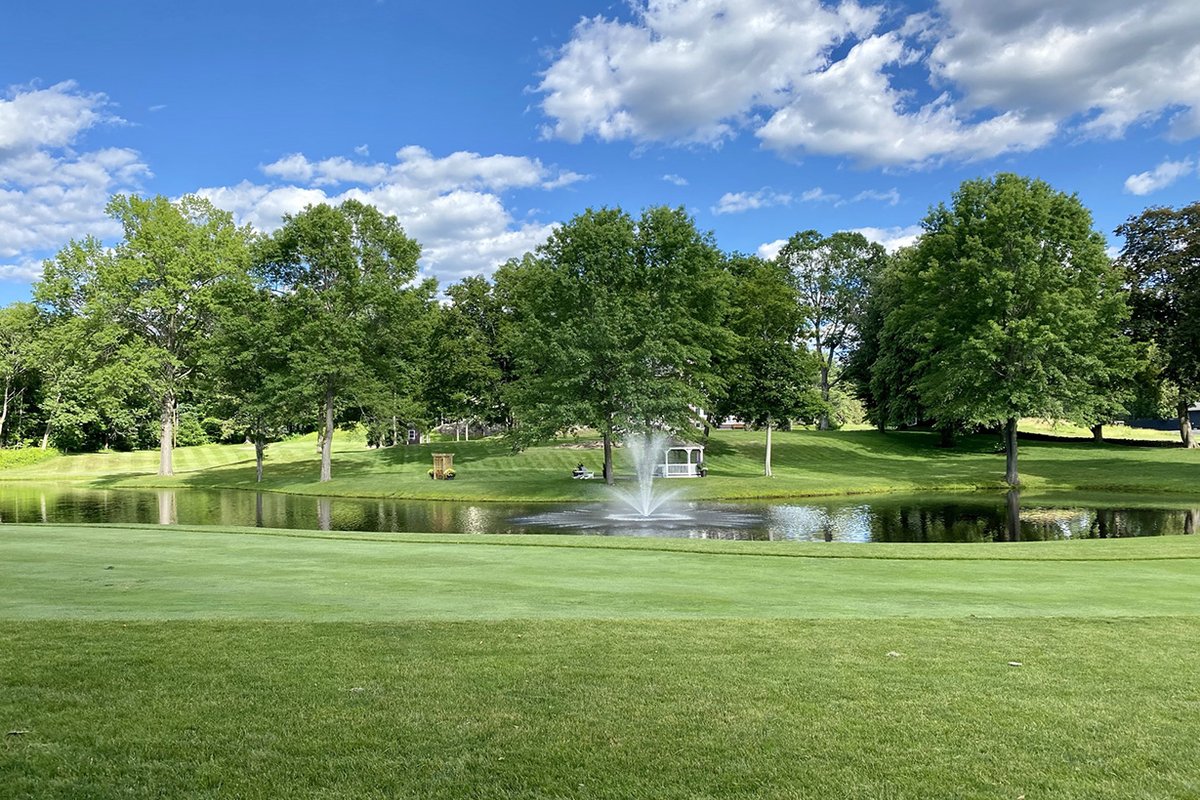 Lush green fairway and manicured putting green at Northfield Golf Course with pond, water fountain, and mature trees under blue sky