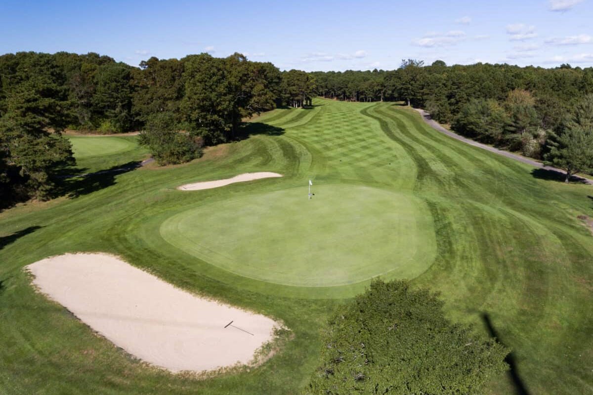 Elevated view of Olde Barnstable Fairgrounds Golf Club green with two bunkers, striped fairway, trees, and blue sky.
