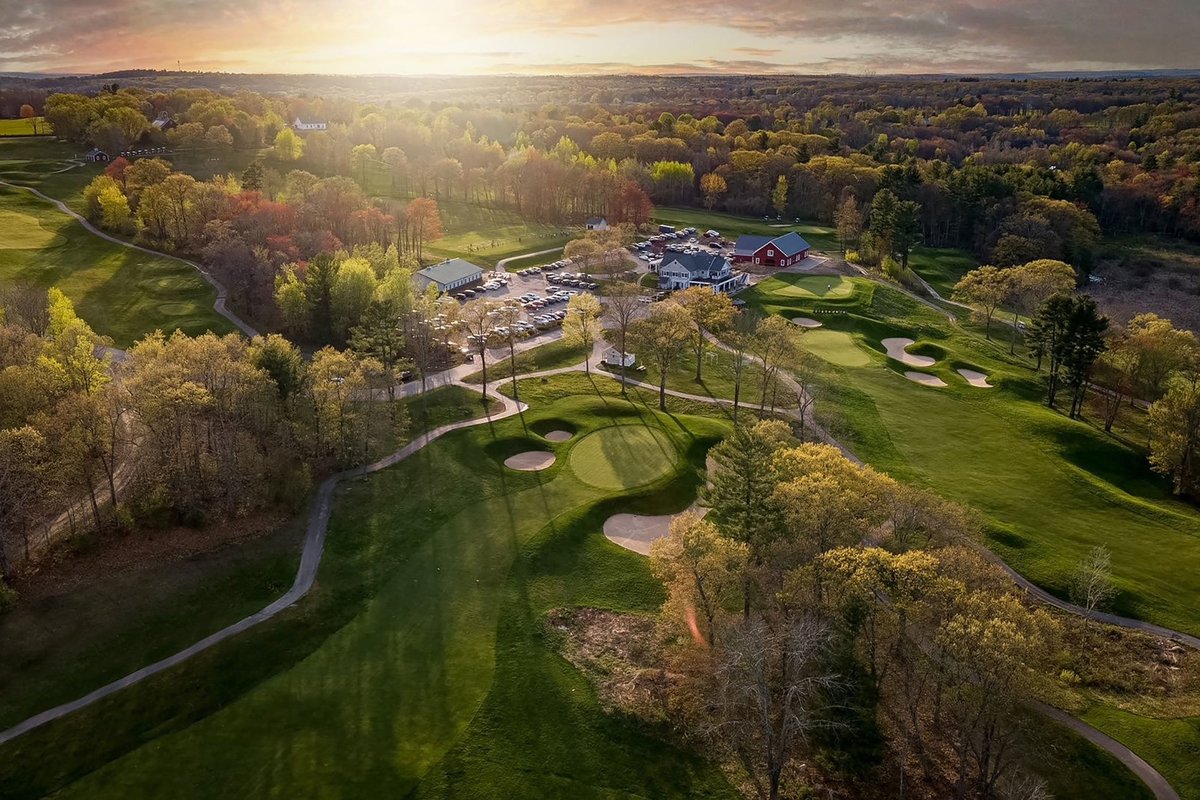Aerial view of Blackstone National Golf Club in Sutton, MA with tree-lined fairways, bunkers, clubhouse, and sunlit sky