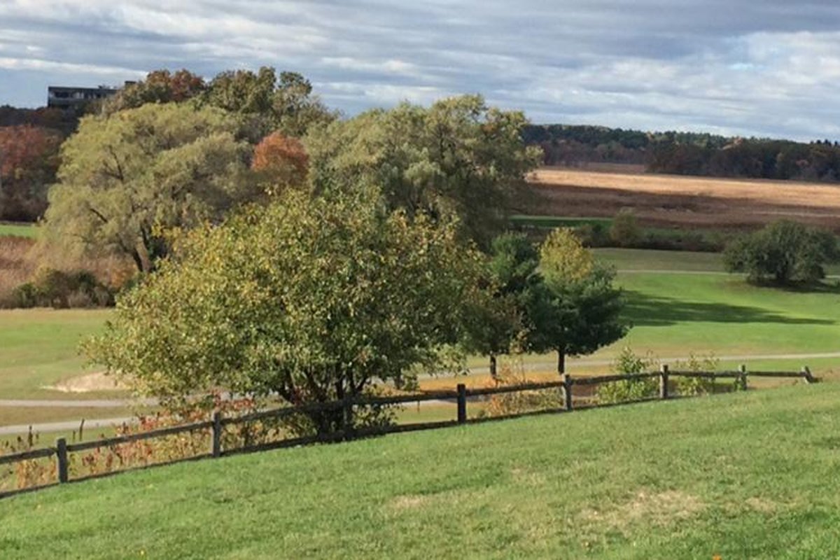 Rolling fairway at Reedy Meadow Golf Course with lush green grass, scattered trees, wooden fence, and distant autumn foliage under a cloudy sky