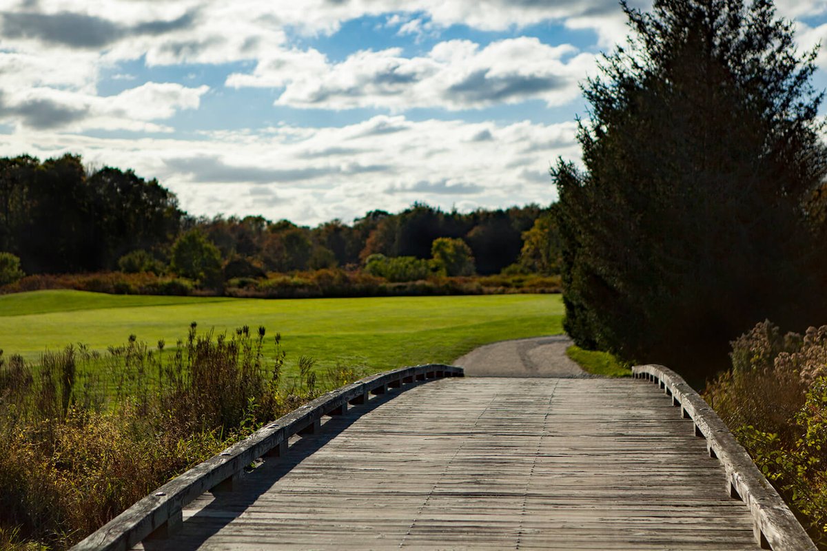 Wooden bridge crosses a path leading to a lush fairway at West Bridgewater Country Club under a partly cloudy sky
