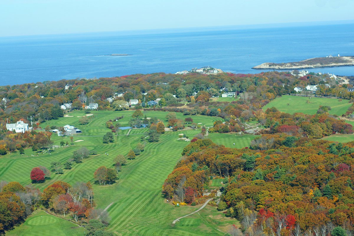 Aerial view of Rockport Golf Club with manicured fairways, autumn trees, scattered homes, and the Atlantic Ocean in the background