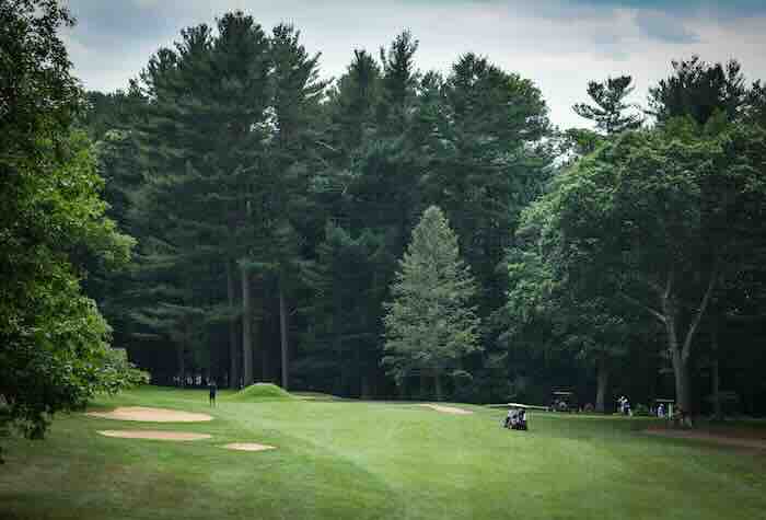 Wide green fairway at Sagamore Spring Golf Club in Lynnfield, MA lined with tall pine trees, bunkers, and golf carts under cloudy sky