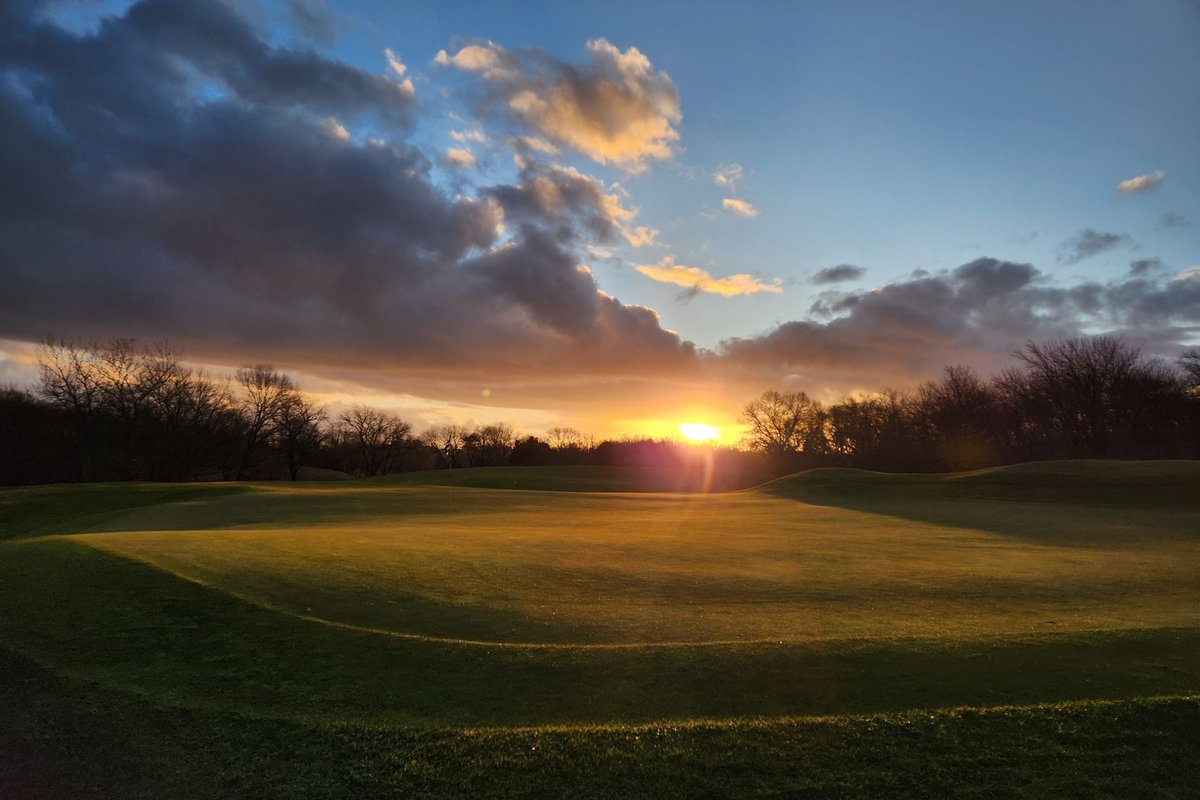 Sunrise over the green at Olde Salem Greens GC in Salem, MA, with long shadows, trees, and dramatic clouds in the sky