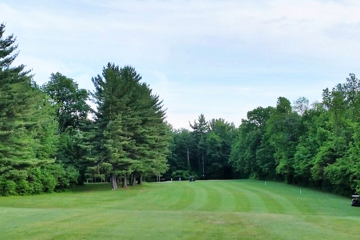 Wide fairway at Settlers Crossing GC in Lunenburg, MA bordered by tall pine trees and lush green rough under a partly cloudy sky