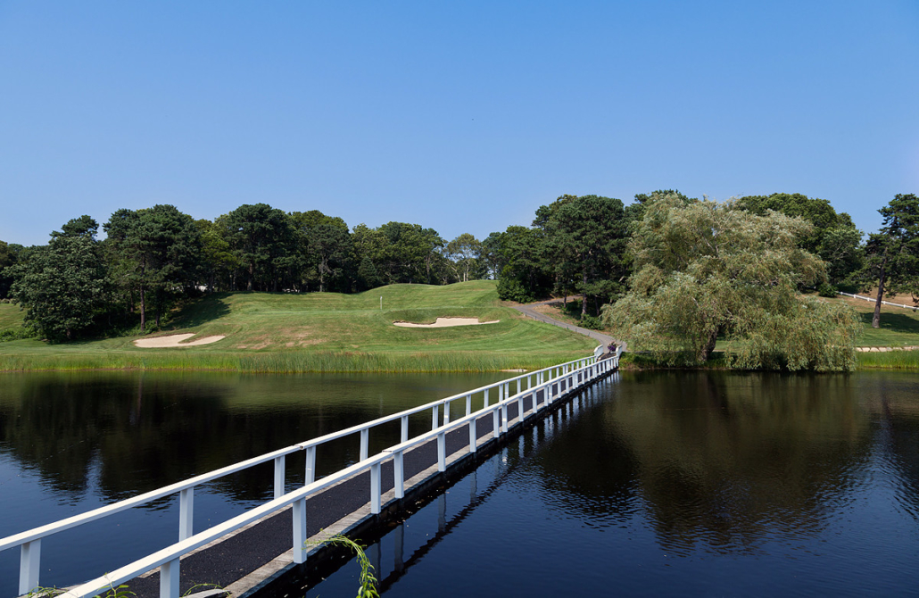 White footbridge crossing a pond toward Blue Rock Golf Course green, flanked by sand bunkers and surrounded by trees under clear blue sky