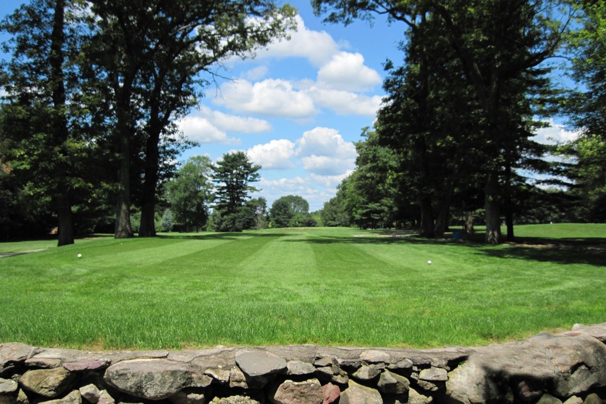 Lush green fairway at Stone E Lea Golf Course in Attleboro, bordered by large trees and a stone wall under a blue sky with clouds