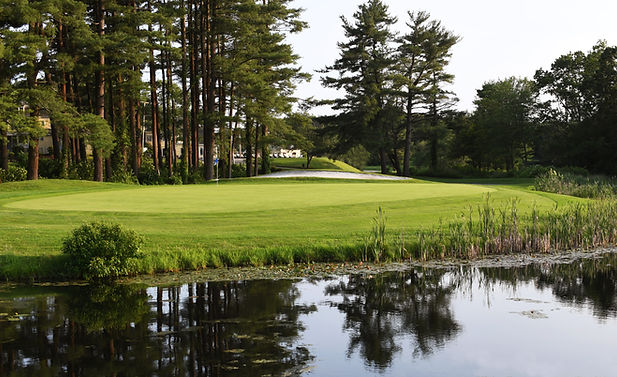 South Course at Stow Acres CC with lush green, pond in foreground, tall pines lining the fairway, and a bright sky above