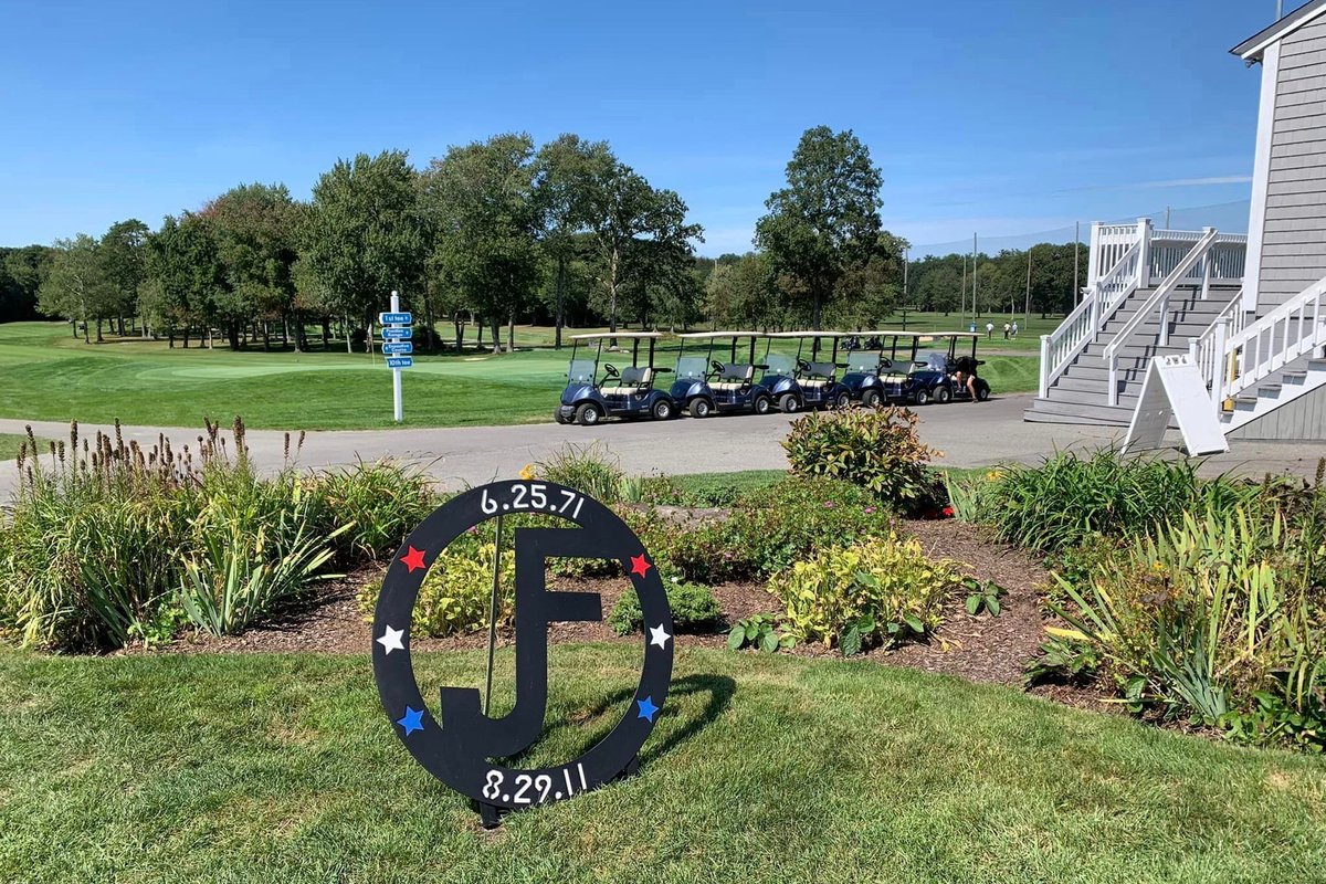 Golf carts lined up near a clubhouse at Swansea Country Club, lush fairway, trees, and blue sky in the background