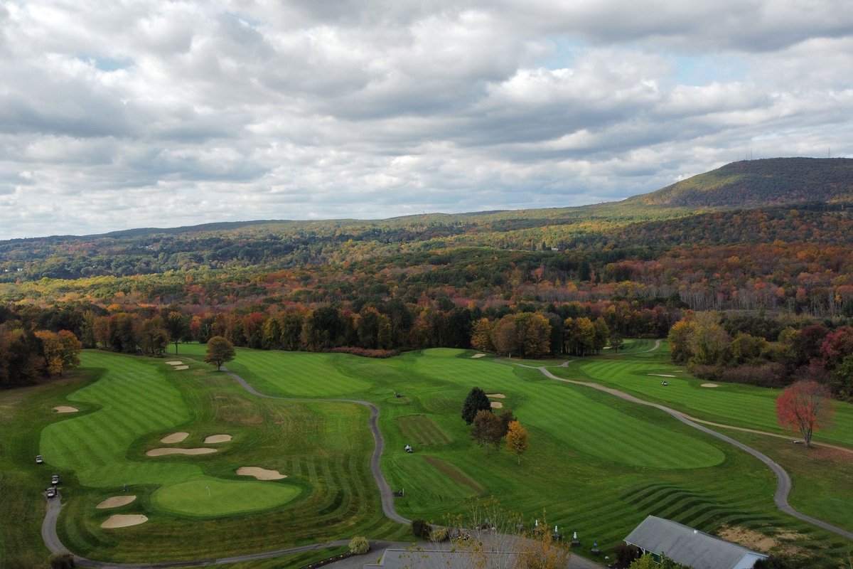 Aerial view of The Ledges Municipal GC in South Hadley, MA showing striped fairways, sand bunkers, trees with autumn colors, and distant hills under a cloudy sky