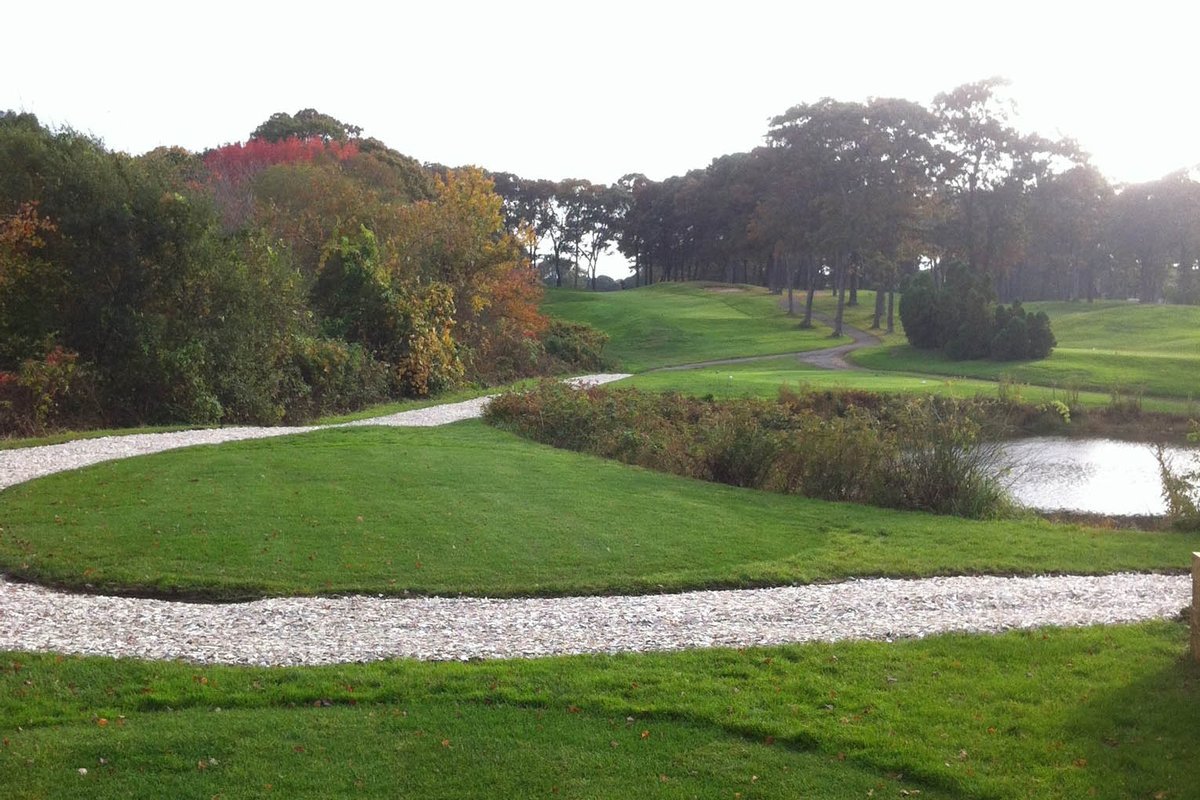 Winding gravel path runs through lush fairways and trees at Twin Brooks Golf Course in Barnstable, MA under a bright sky.