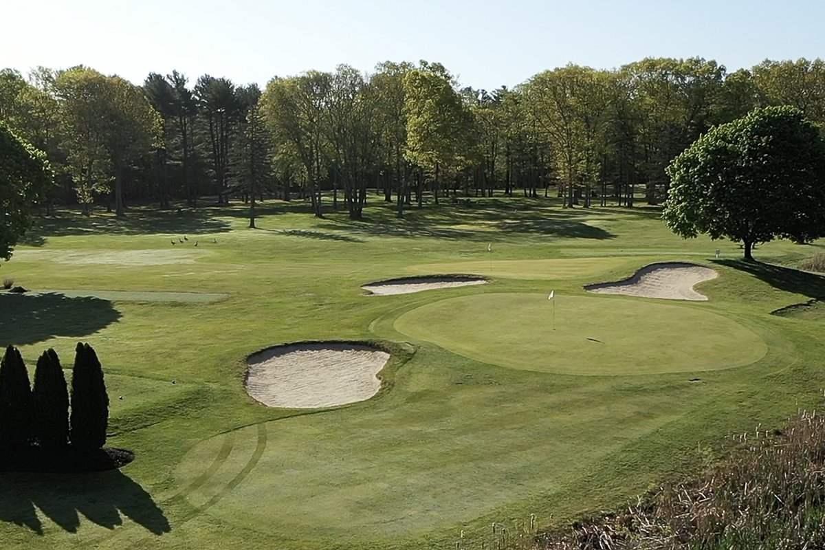 Sunny day view of MGA Links at Mamantapett’s green with three sand bunkers, fairway, scattered trees, and blue sky in Norton, MA