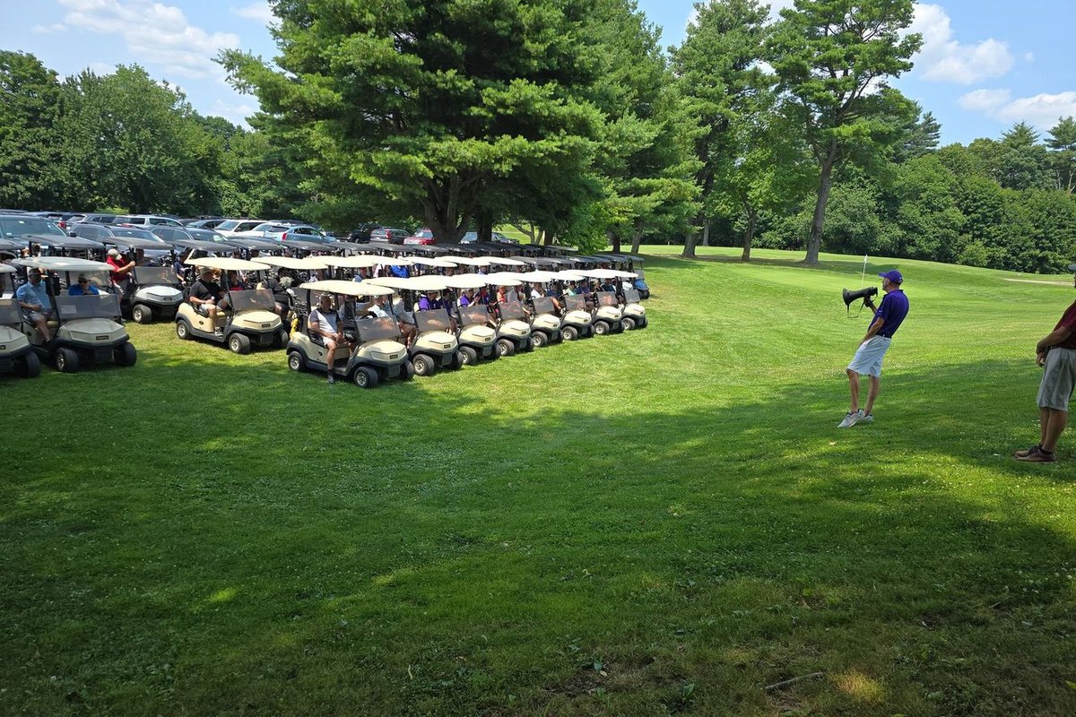 Dozens of golf carts line up on lush green grass at Wenham Country Club as a man with a megaphone addresses golfers under tall trees.