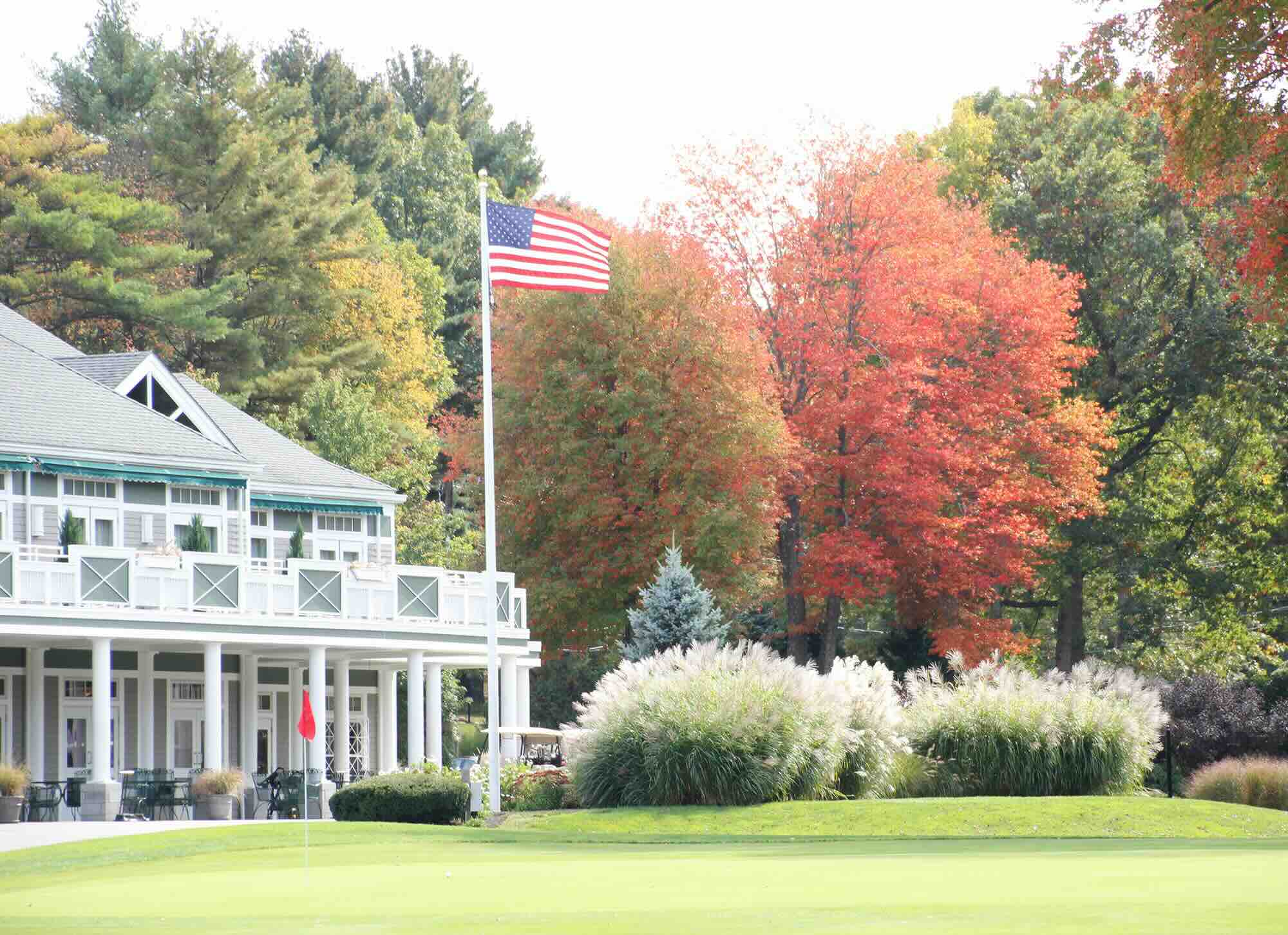 Clubhouse at Brookmeadow Country Club with American flag, lush green putting green, colorful autumn trees, and manicured shrubs