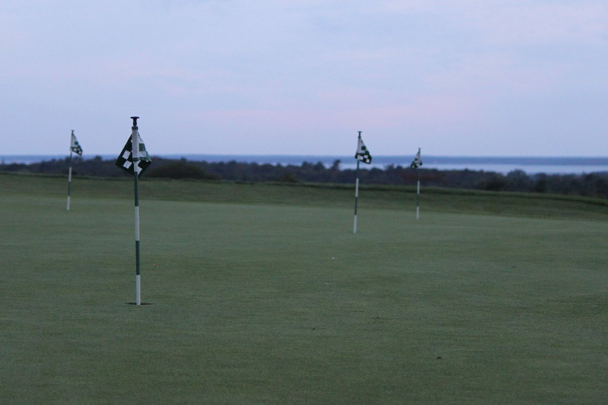 Multiple putting green flags on the practice green at The Brookside Club in Bourne, MA, with trees and water in the background