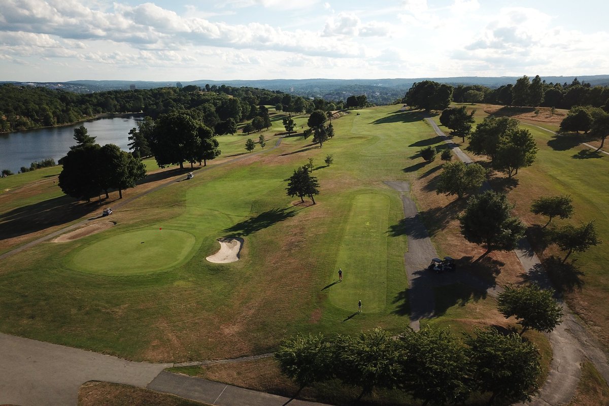 Aerial view of Green Hill Municipal GC in Worcester, MA featuring fairways, greens, sand bunkers, scattered trees, and a lakeside backdrop