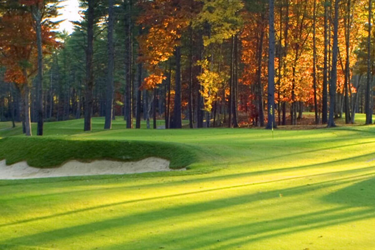 Sunlight casts long shadows across a green and sand bunker at Butter Brook Golf Club, surrounded by autumn trees in Westford, MA