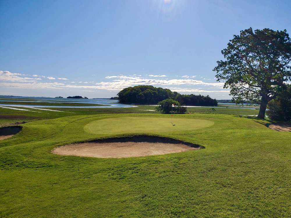 Cape Ann Golf Course green surrounded by sand bunkers, overlooking marshland, water, and trees under a clear blue sky